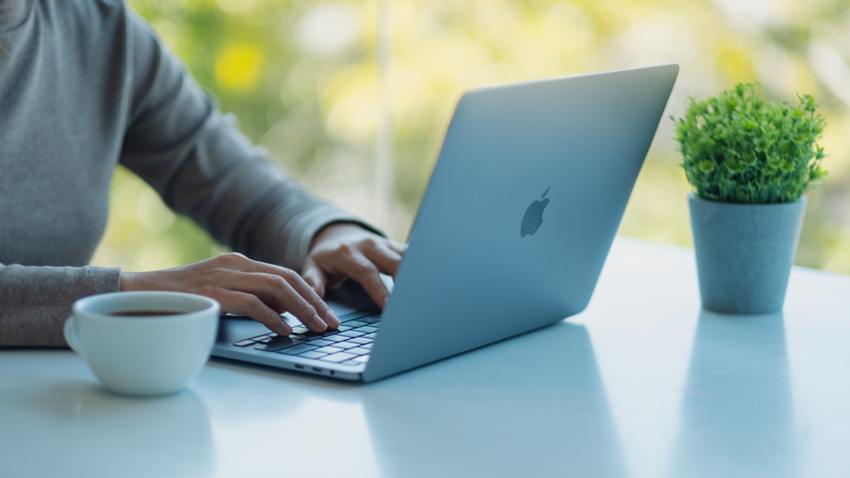 Woman at table with coffee cup and plant using her Apple Macbook after learning how to recover deleted files on Mac.